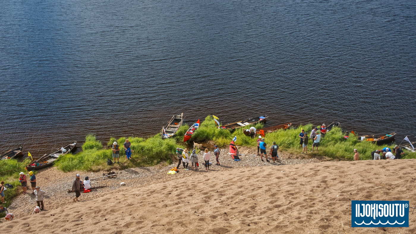 Kuvassa näkymä ylhäältä Jurmun hiekkatörmältä. Iijokisoudun osanottajia, veneitä ja kajakkeja on rannassa.