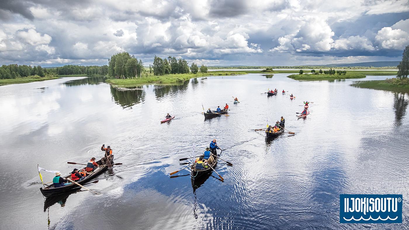 A photograph from the fourth rowing day of Iijokisoutu. A view from a bridge by the Sotkajrvi lake. The boats and kayaks slide across the surface of the like in soft light, as the sky is reflected in the water.